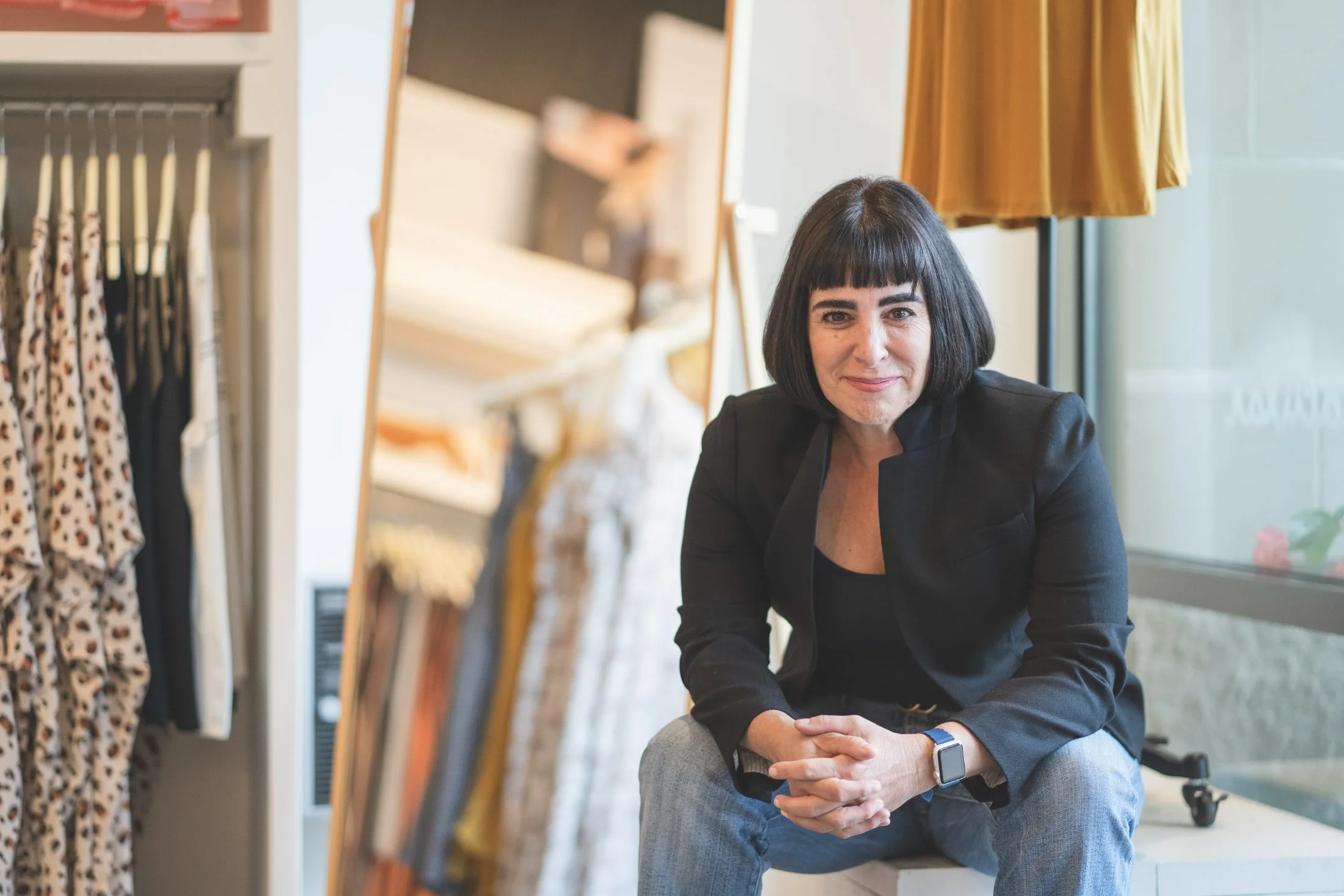 Rachel Williamson seated on a fixture inside a clothing store, hands clasped, looking at the camera