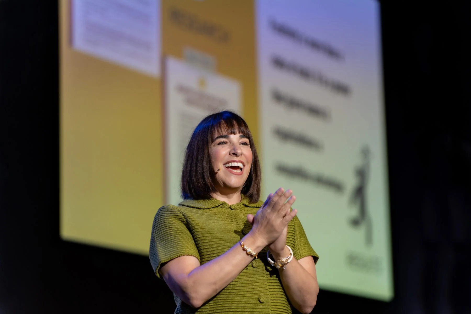Rachel Williamson on stage mid-keynote, applauding, with presentation slides behind her