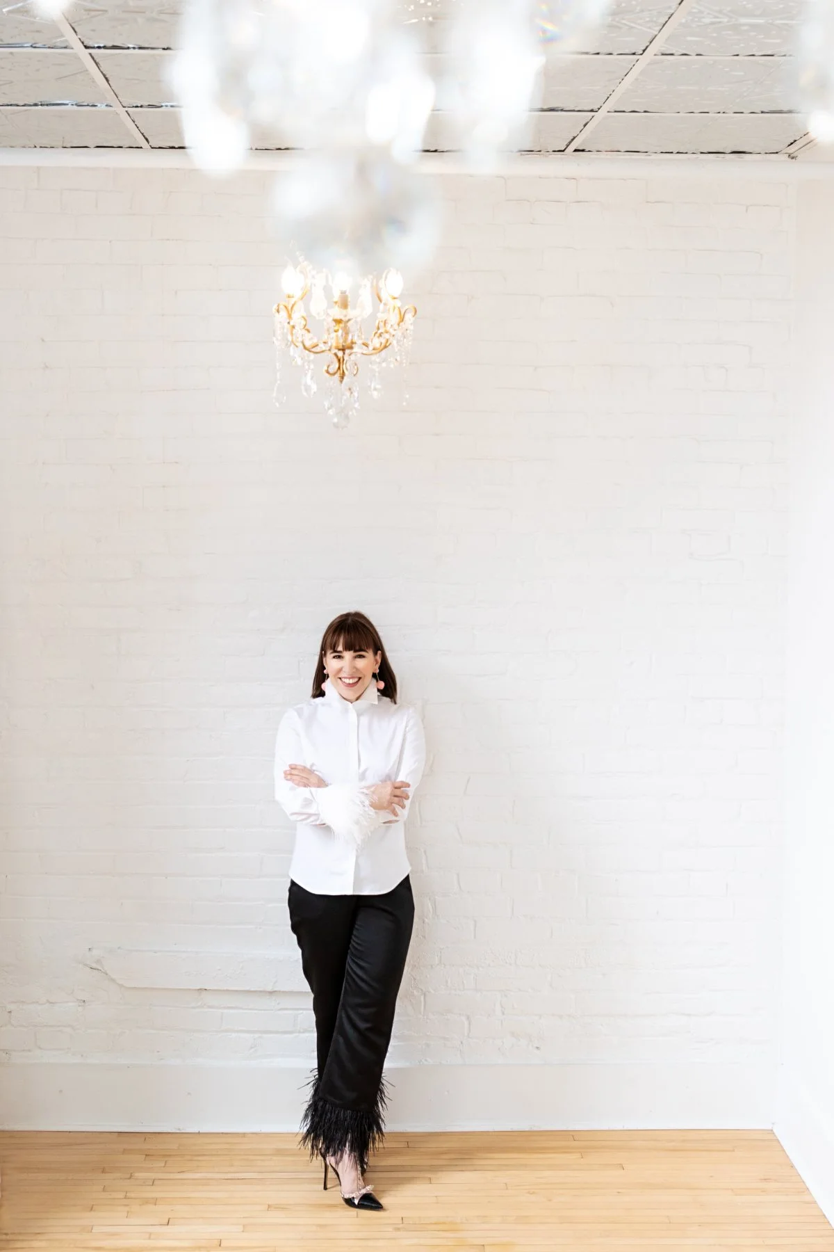 Rachel Williamson standing with arms crossed against a white brick wall beneath a chandelier